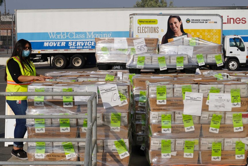 A woman wearing a mask and high vis looks at piles of boxes with a note of Ballots only on green paper