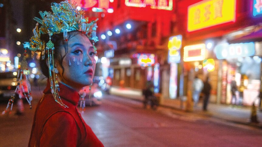Still from Rainbow Chan's 'Seven Sisters' music video. She is dressed in red and standing on a street at night, lit in neon.