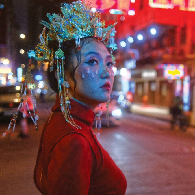 Still from Rainbow Chan's 'Seven Sisters' music video. She is dressed in red and standing on a street at night, lit in neon.