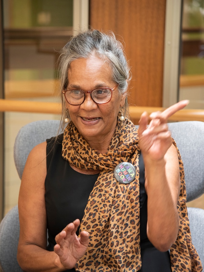 Indigenous woman Ms Moreton sits in the library, wearing glass and a leopard scarf pointing to the distance.