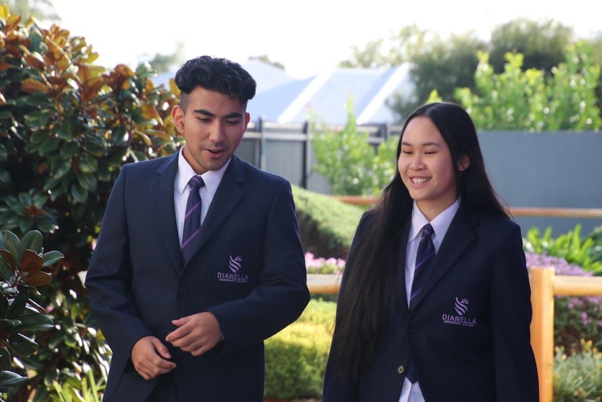 John and Kathy wearing formal school uniform, standing outside in sunshine near trees and shrubs, talking to each other
