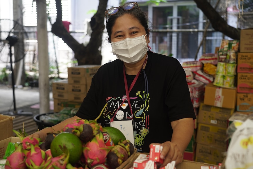 A Thai woman in a mask stands behind a case of fruit 