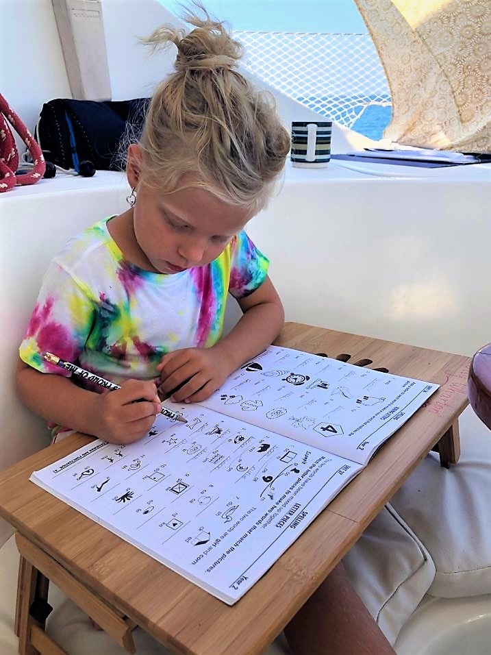 A young girl in a colourful t-shirt writes in an illustrated exercise book at a small table on a boat.