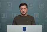 Volodymyr Zelenskyy stands at a podium in front of a Ukrainian flag with a backdrop that says "the office of the president". 