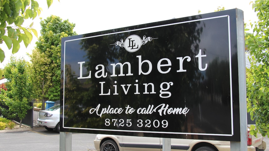 A black sign with white writing saying 'Lambert Living, a place to call home' with trees in the background