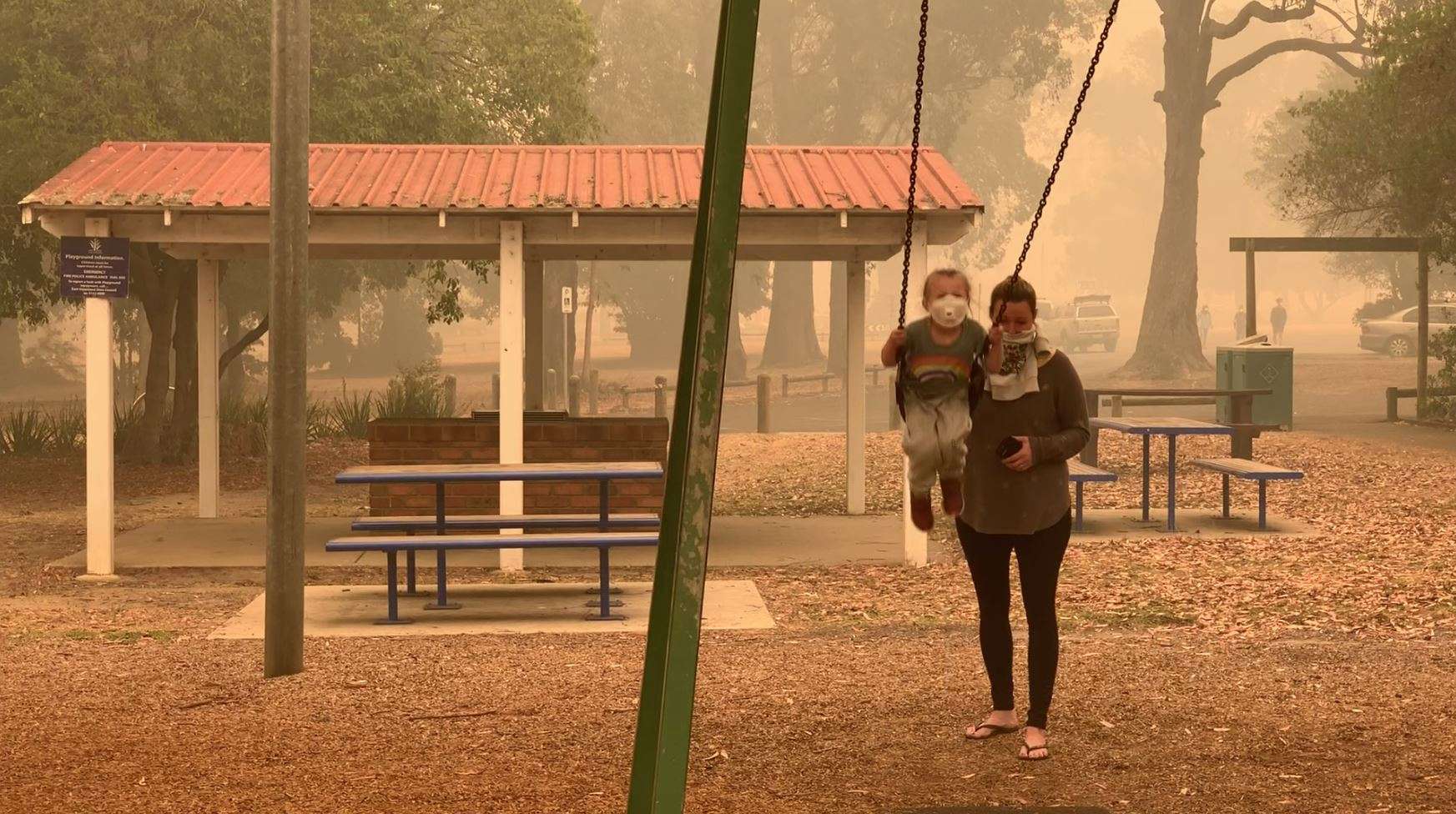 A woman pushes a child on a swing, both of them wearing dust masks, as an orange haze hangs in the air in Mallacoota.
