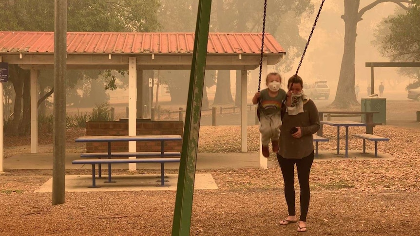 A woman pushes a child on a swing, both of them wearing dust masks, as an orange haze hangs in the air in Mallacoota.