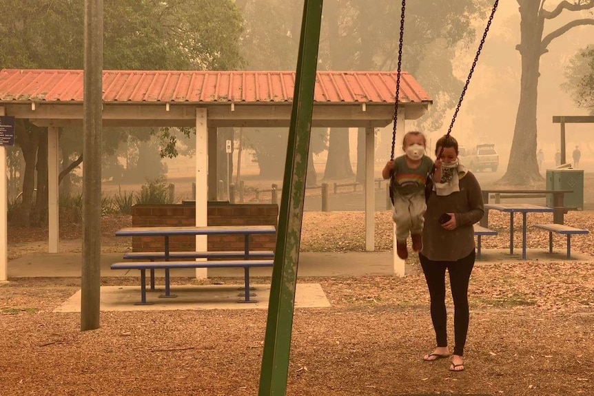 A woman pushes a child on a swing, both of them wearing dust masks, as an orange haze hangs in the air in Mallacoota.