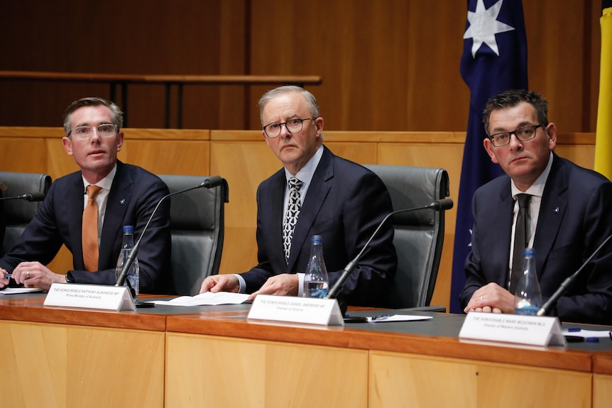 Трима мъже в костюми седят покрай бюро с микрофони