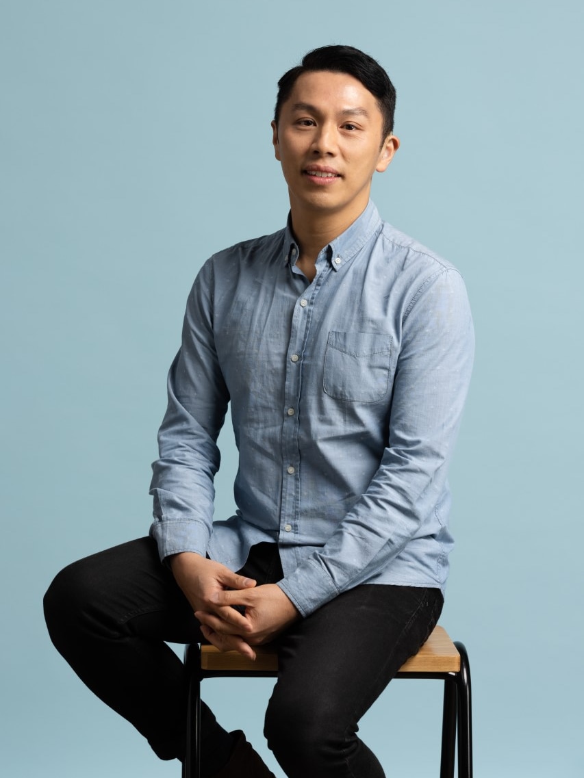 A man in a blue shirt and black pants sits on a still in front of a blue background.