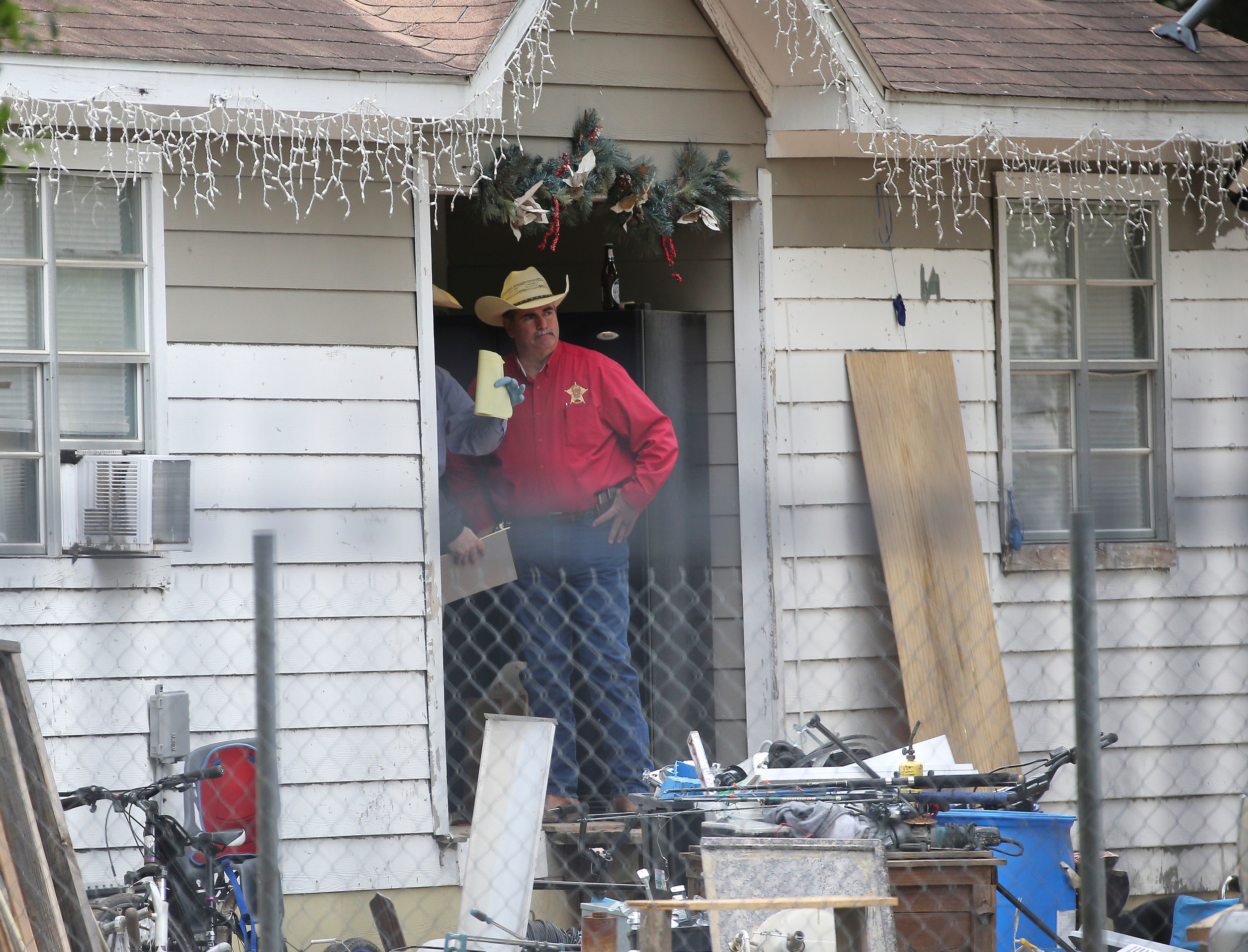 A sheriff in a cowboy hat and red shirt looks out from the doorway of a white weatherboard home with hands on hips.