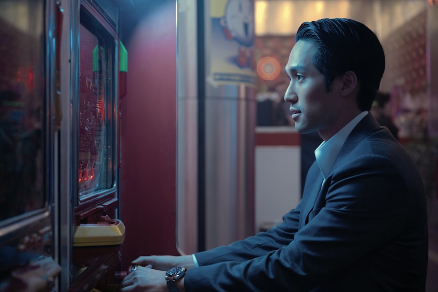 A Korean man in a suit sits in profile, he is playing pachinko on a machine in a pachinko parlour.