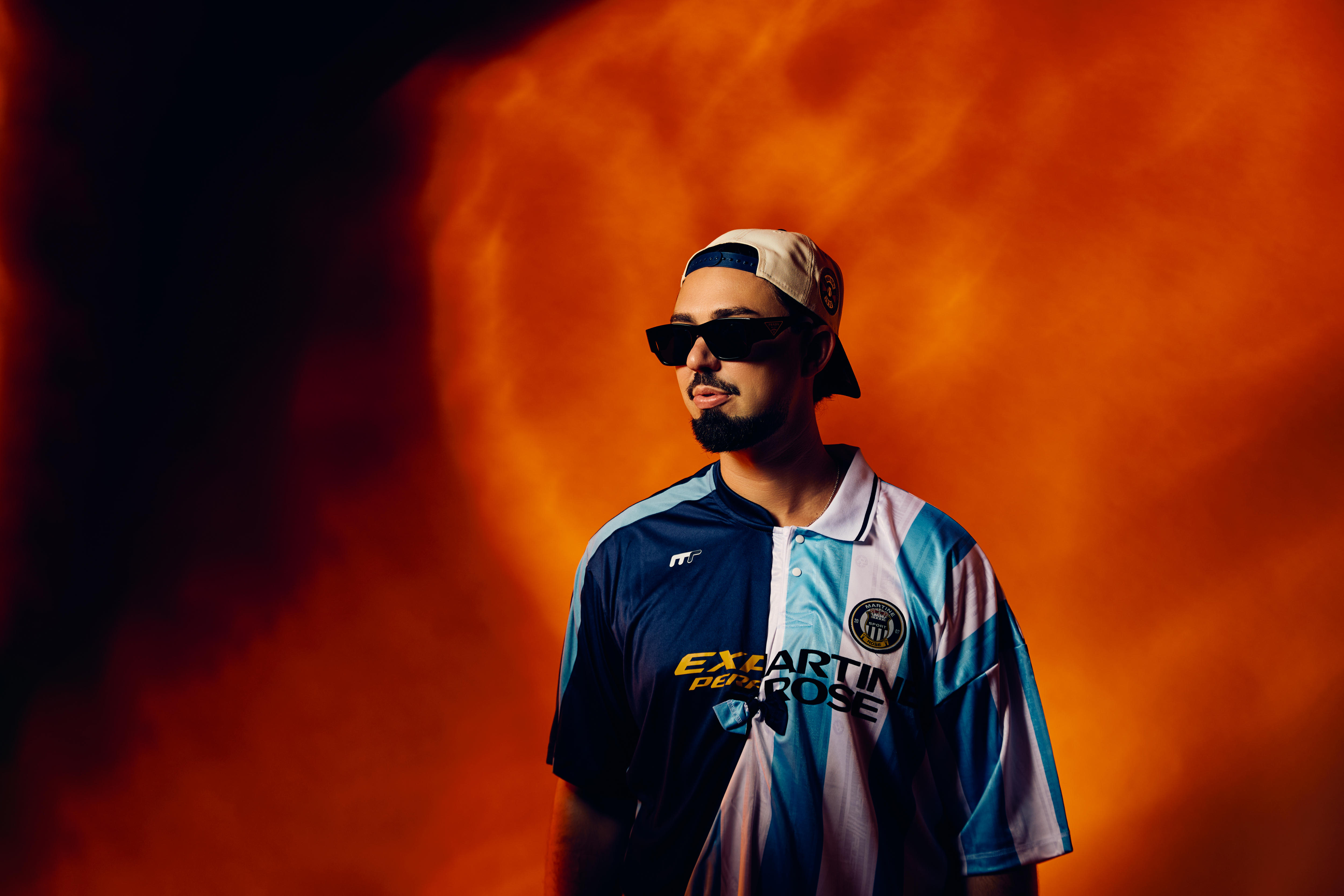 A man in a backwards white cap, black sunglasses and a blue and white jersey stands in front of an orange background