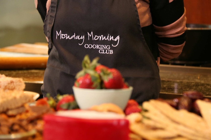 A women wears an apron with the words Monday Morning Cooking Club stitched on it.