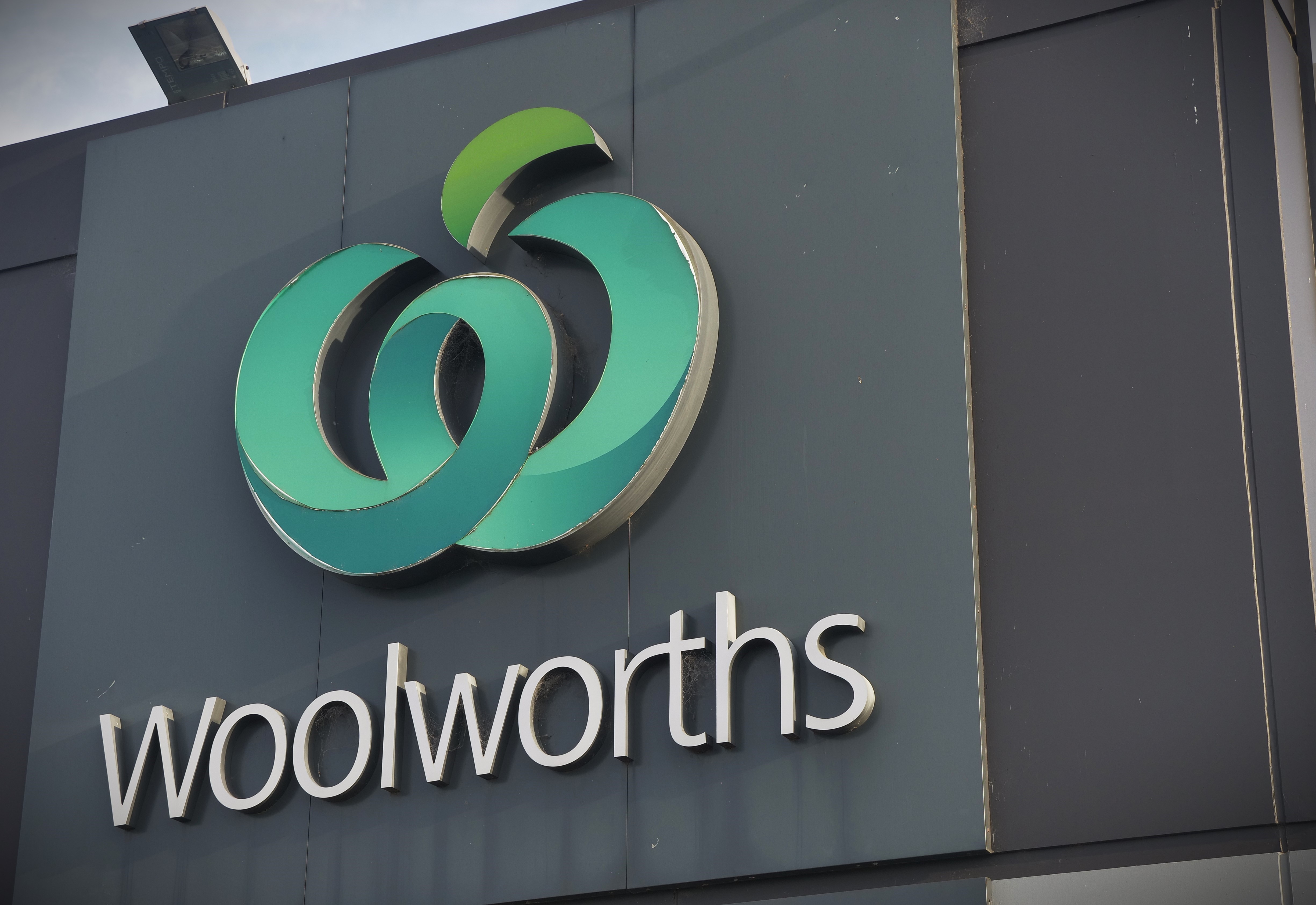 Um close da placa e do logotipo da Woolworths fora de uma loja. O logotipo é um 'w' verde em forma de maçã. 