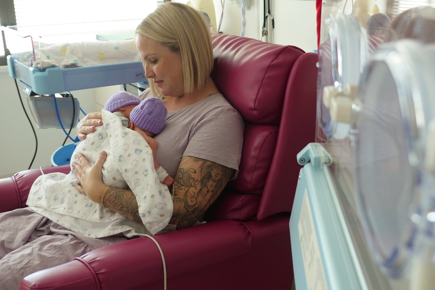 Tamara sitting in a red chair holding Tide and Kove on her chest, incubator in the foreground.