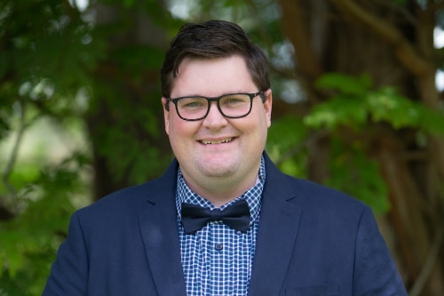 A man with short dark hair and black glasses smiles. He is wearing a navy suit with a blue cheque shirt and bow tie.