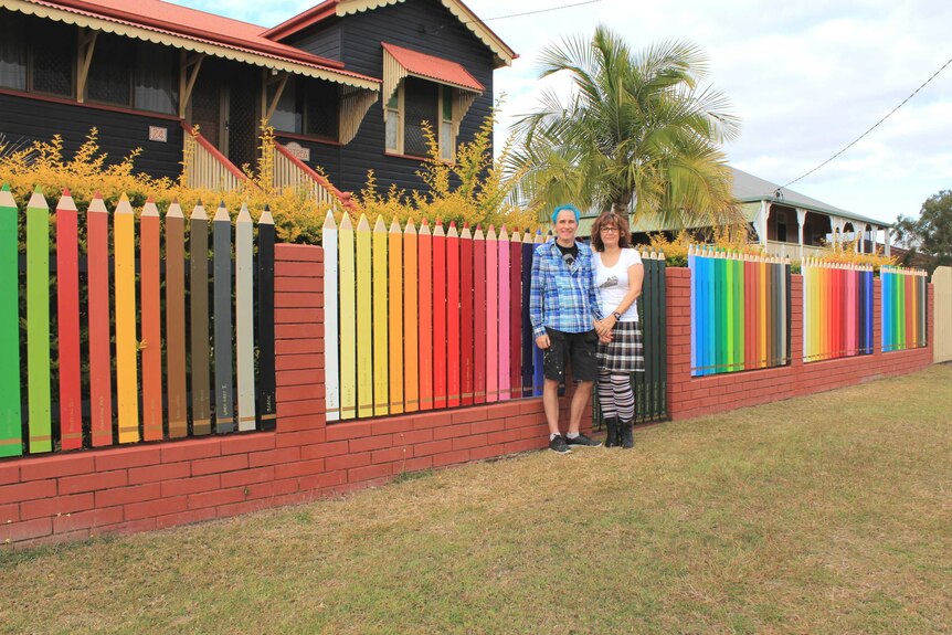 Phil and Marlies Oakley stand in front of a wooden fence made to look like a set of 108 different coloured pencils.