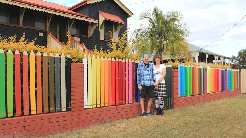 Phil and Marlies Oakley stand in front of a wooden fence made to look like a set of 108 different coloured pencils.