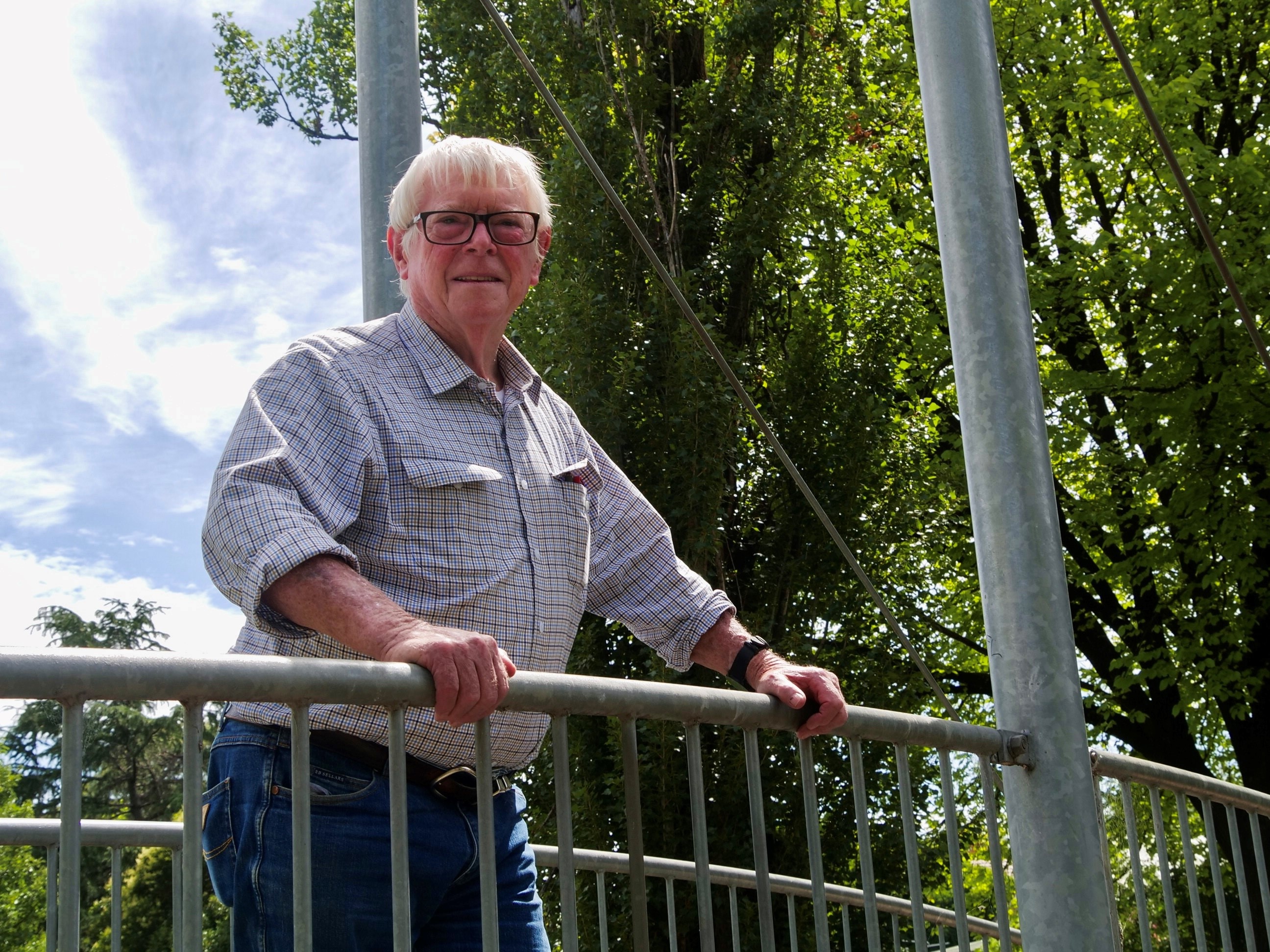 A man in a plaid shirt stands on a bridge in a park.