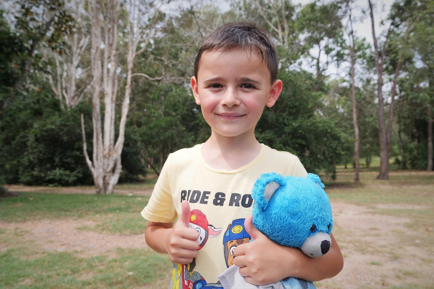 Small boy gives a thumbs up and holds a blue teddy bear.
