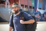 A bearded man, with blue eyes, carrying a backpack walking in front of a cargo ship, wears blue t-shirt, hat, looks seriously.