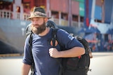 A bearded man, with blue eyes, carrying a backpack walking in front of a cargo ship, wears blue t-shirt, hat, looks seriously.