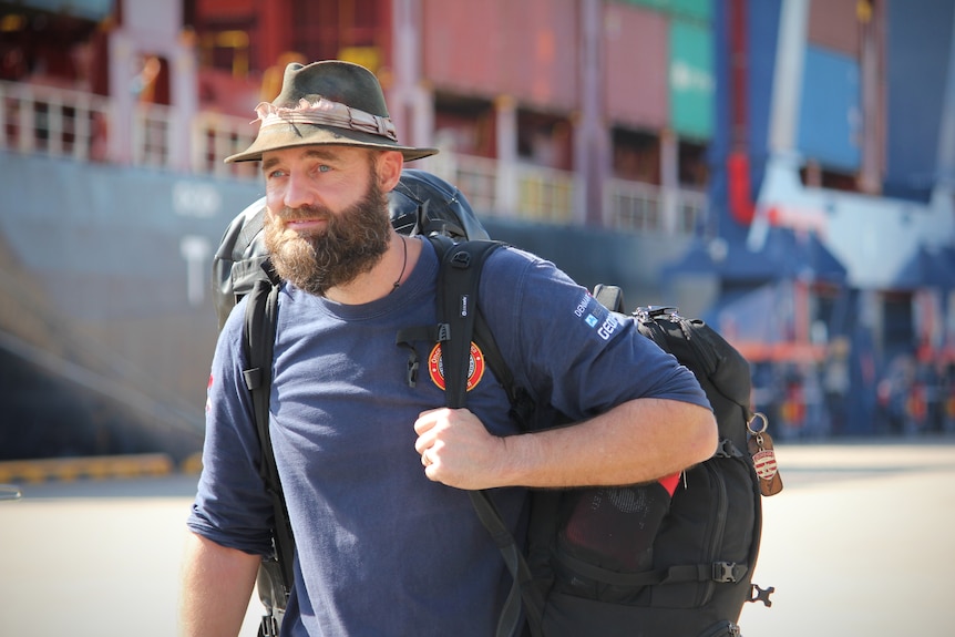 A man carrying a backpack walking in front of a cargo ship