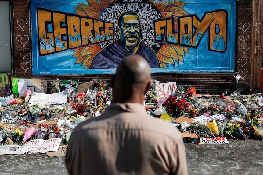 A black man stands in front of a mural of George Floyd