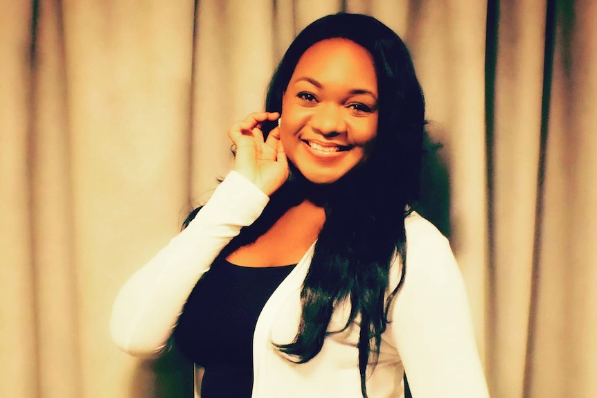 A beautiful black woman with long dark hair, holding a hand to her ear, wearing a black top with a white cardigan