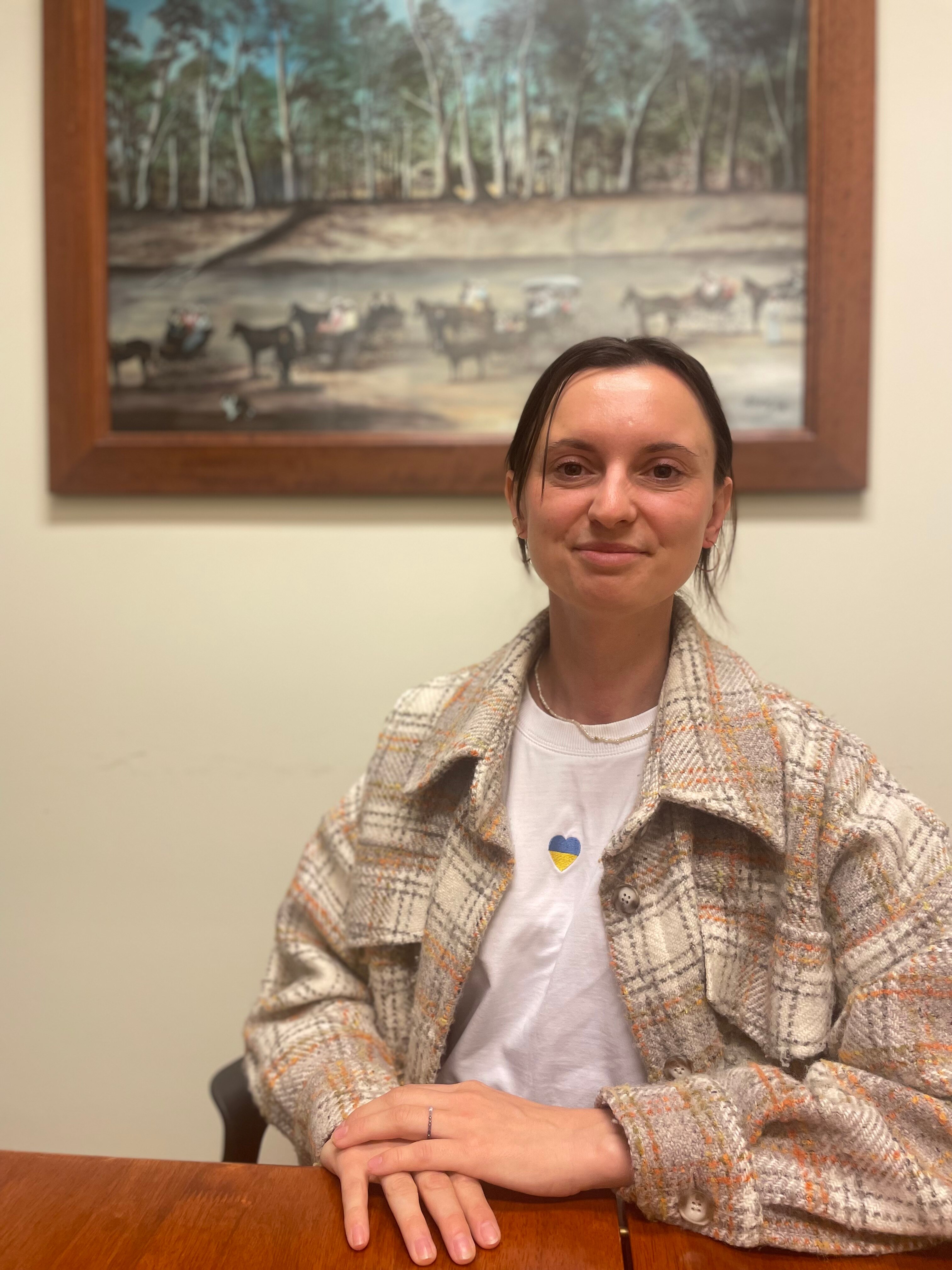 A smiling woman sitting down in front of a painting, wears cream check court, hair tied back.