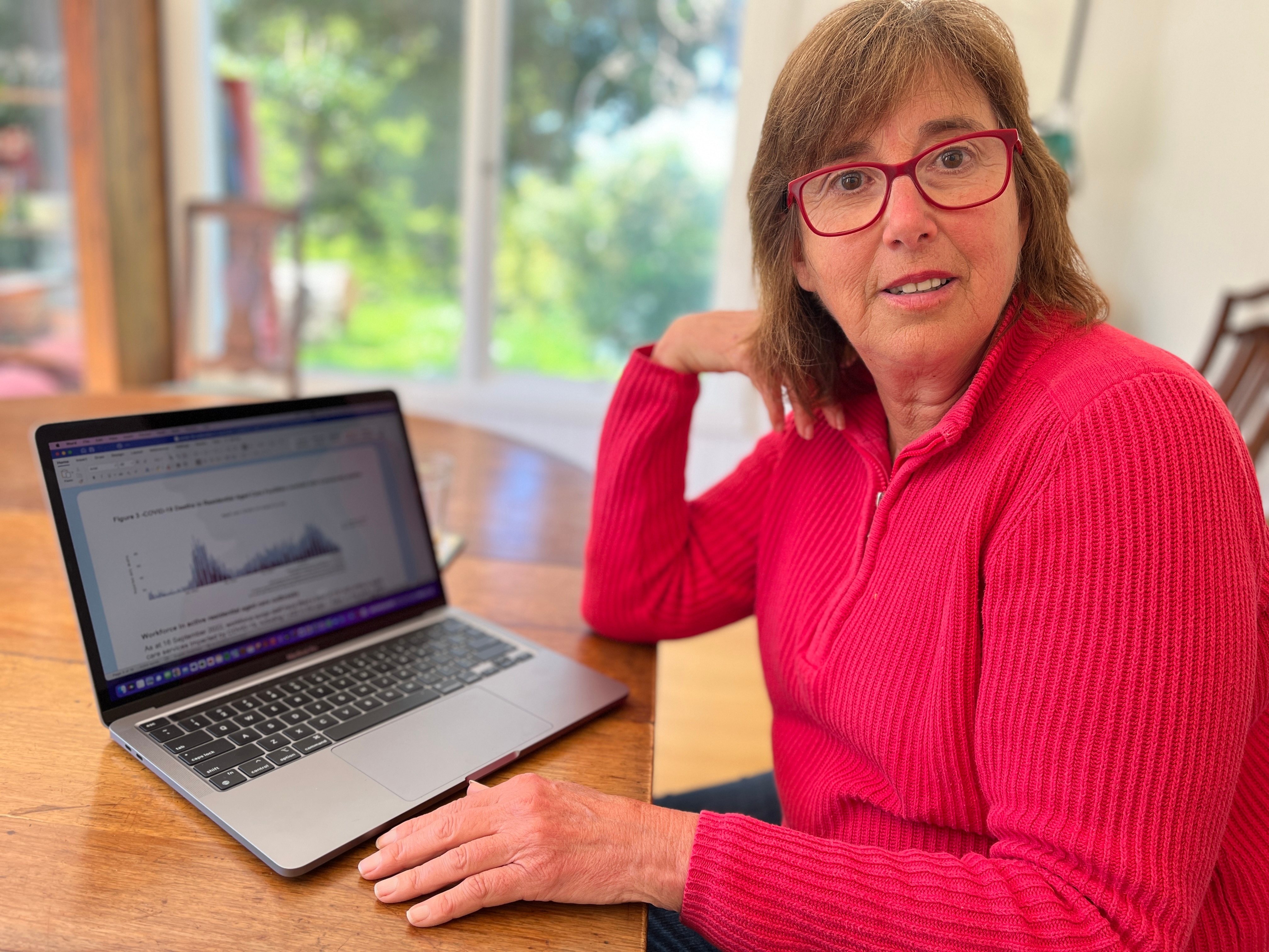 A woman sitting at a table with a laptop. She has red glasses and a pink top