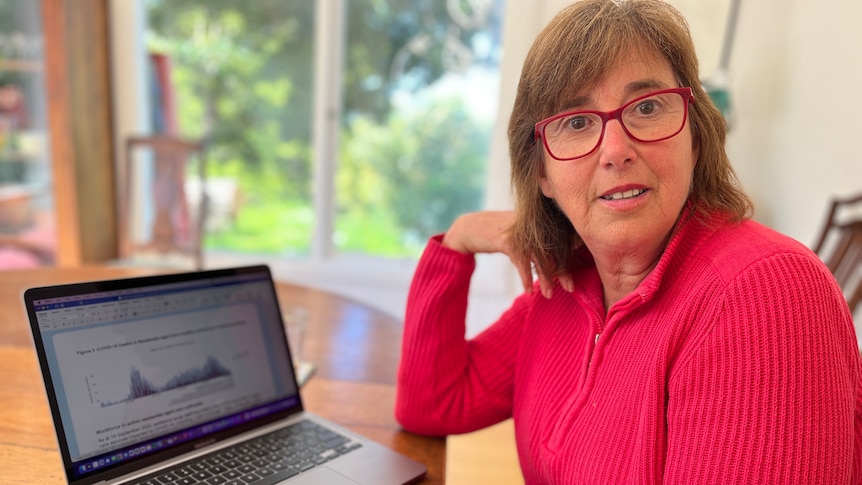 A woman sitting at a table with a laptop. She has red glasses and a pink top