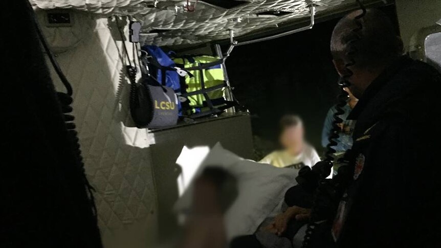 Blurred out, dark image of a boy lying against a pillow with paramedic equipment and emergency services standing in background.