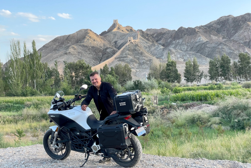 A man poses with a motorcycle in front of mountains with a castle wall running along them