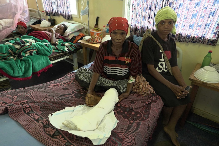 An elderly woman with a cast on her leg sits in a hospital bed as another lies in the next bed.