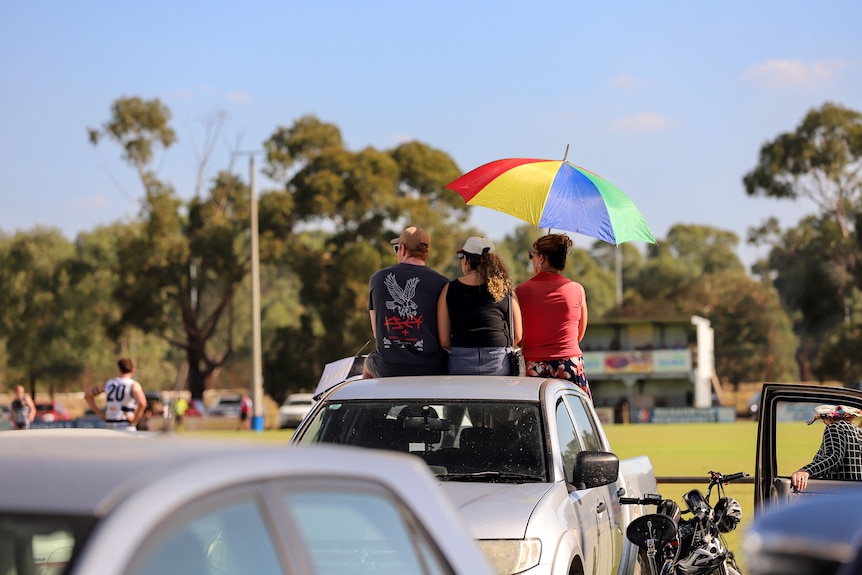 Three people sit on top of ute car tray under rainbow coloured umbrella, watching a country football match