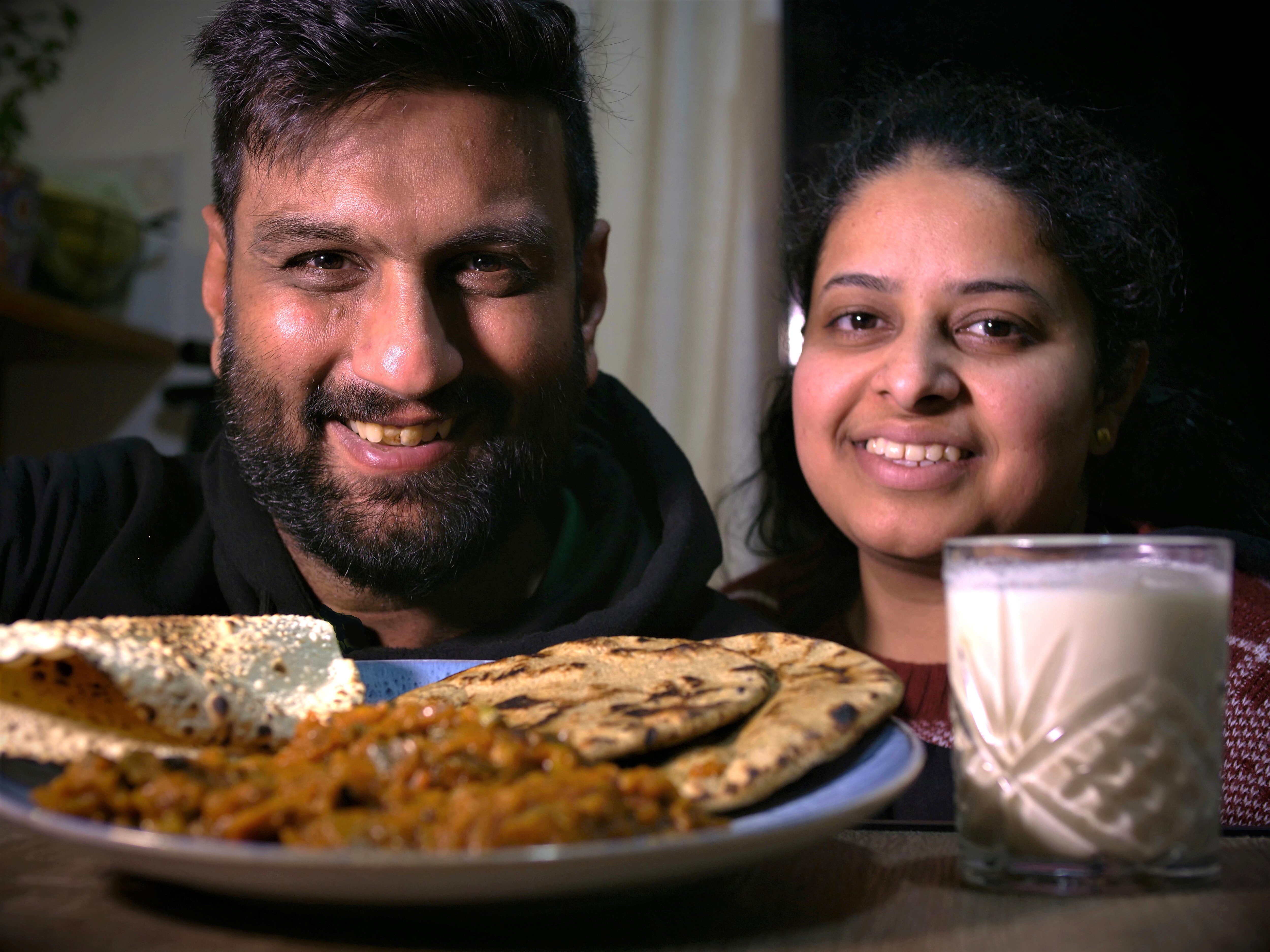 An Indian man and woman smiling, with a plate of Indian food in the foreground.