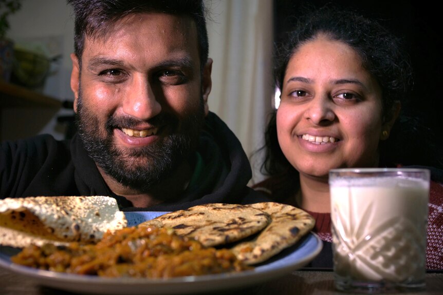 An Indian man and woman smiling, with a plate of Indian food in the foreground.
