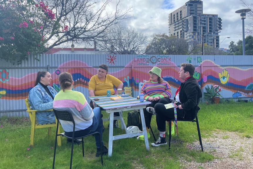 Five young adults sitting around a table talking with a colourful mural behind them.
