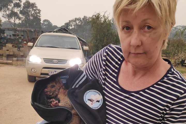 A woman with short blonde hair looks sadly at the camera, holding a possum in her arms.