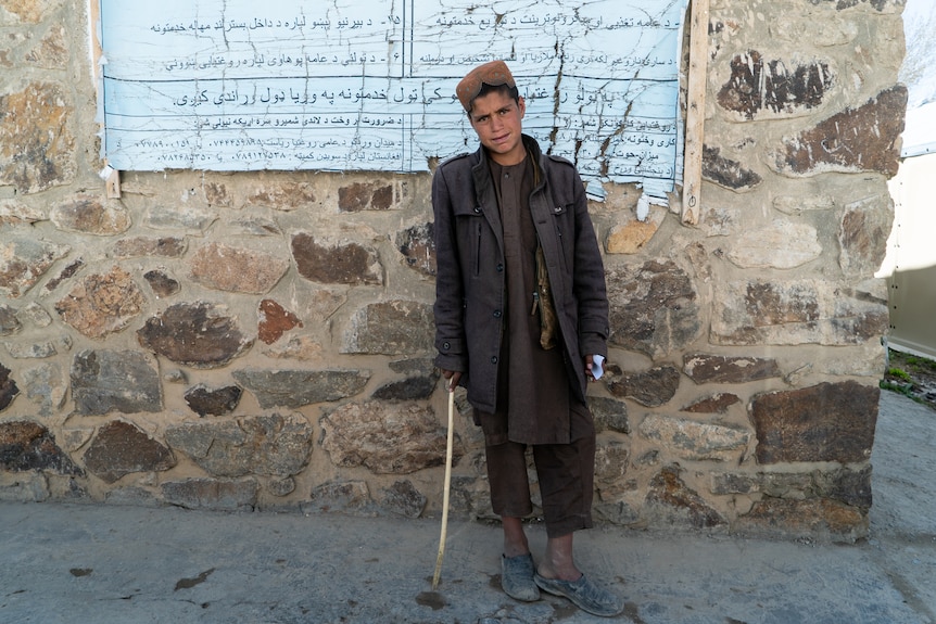 A young boy stands against a stone wall wearing dark robes and leaning on a stick