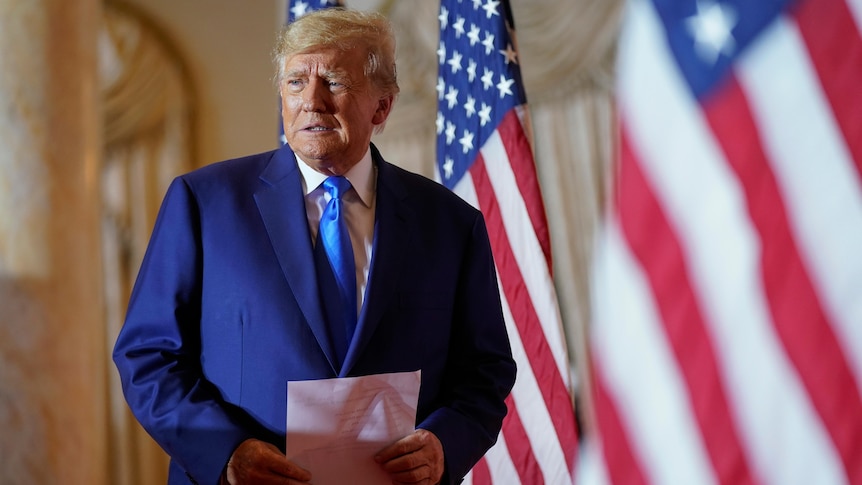 Donald Trump holds documents as he looks over his shoulder while dressed in a suit and standing in front of US flags.