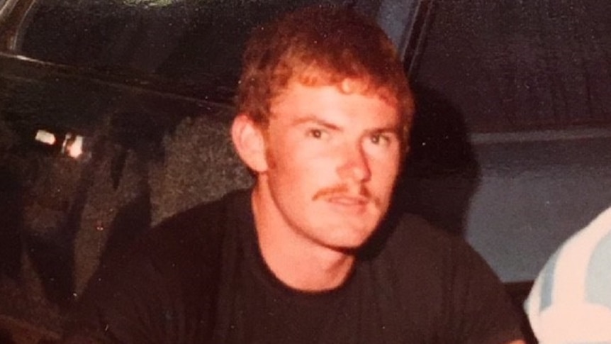 A red headed man with a moustache dressed in black is squatting behind a vehicle