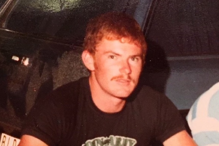 A red headed man with a moustache dressed in black is squatting behind a vehicle