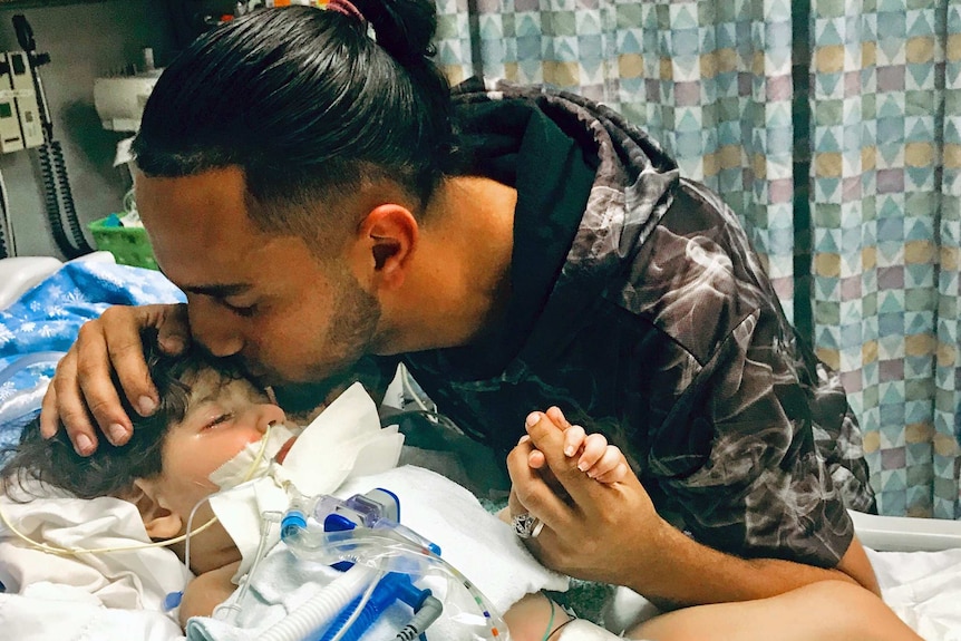 a man kisses the forehead of his son who is in a hospital bed