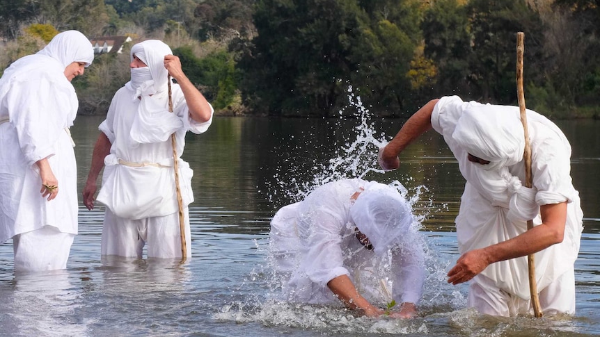 Mandaean man being baptised in river by priest, with two men standing behind. All wear white robes.