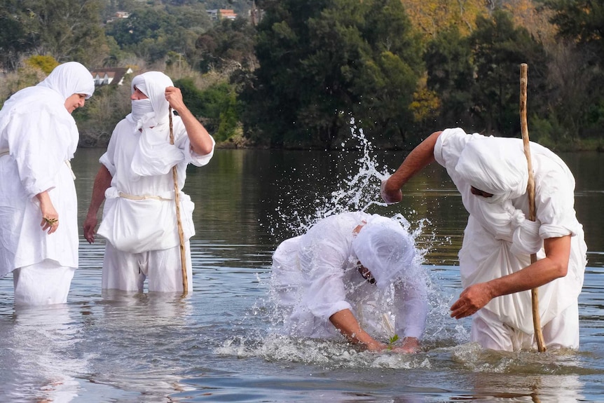 Mandaean man being baptised in river by priest, with two men standing behind. All wear white robes.