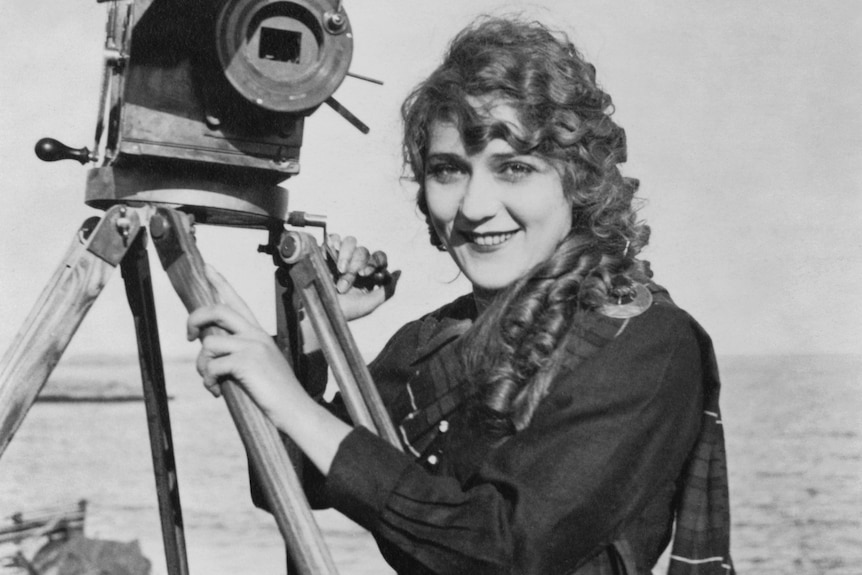 Black and white image of Mary Pickford, smiling widely, holding a large video camera next to her face.