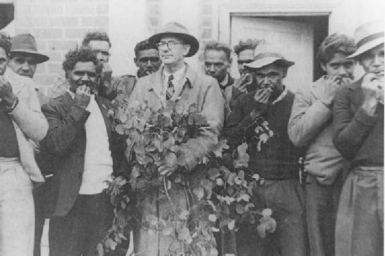 A black and white photo of a group of Indigenous men playing gum leaves, a white man in trench coat, hat, holds bunch of leaves.